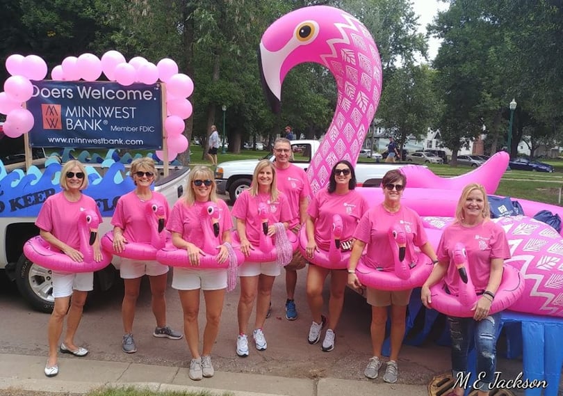 Minnwest team in parade with a giant flamingo float wearing flamingo floaties a